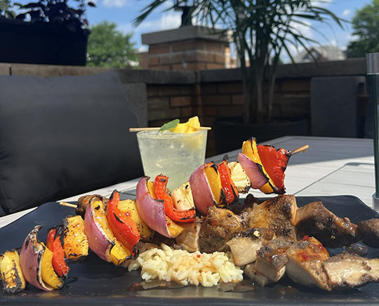 Grilled chicken and vegetable skewers served with rice, alongside a refreshing cocktail, against a sunny outdoor setting.