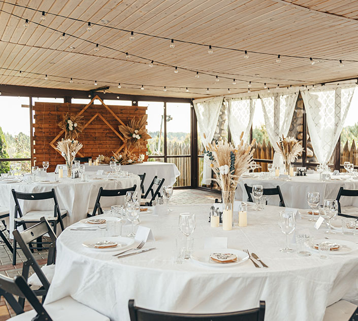 Elegant indoor reception setup with white tablecloths, floral centerpieces, and string lights, featuring a rustic wooden backdrop.
