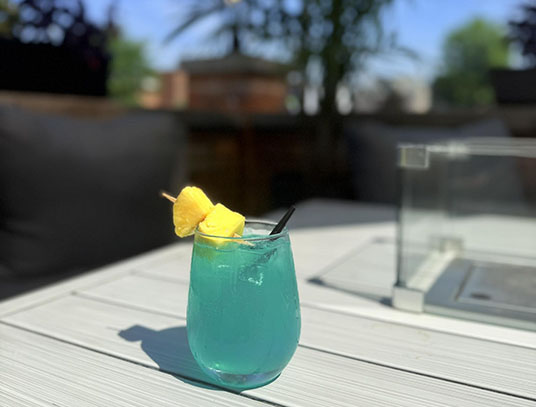 A vibrant blue cocktail garnished with pineapple on a skewer, resting on a sunlit outdoor table with black cushions in the background.