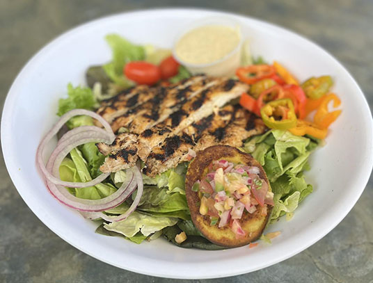 Grilled fish topped salad with lettuce, cherry tomatoes, sliced onions, colorful peppers, and a side of sauce in a bowl.