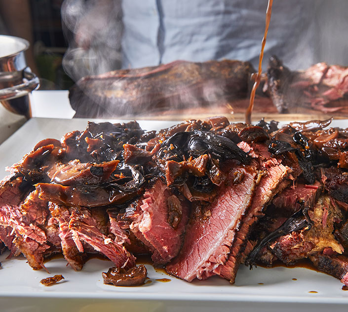 A close-up of tender, sliced beef brisket topped with caramelized onions, steaming and drizzled with sauce on a white platter.