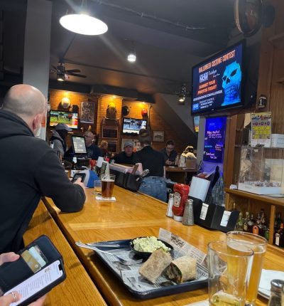 People sitting at a bar, with a television on the wall and a laptop open on the counter in front of them.