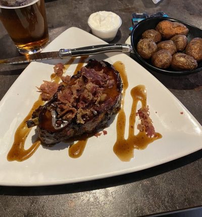 A plate featuring a juicy steak, seasoned potatoes, and a glass of beer beside it.