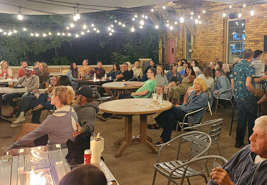 A lively outdoor gathering is illuminated by string lights, with guests seated around tables and enjoying the evening atmosphere.